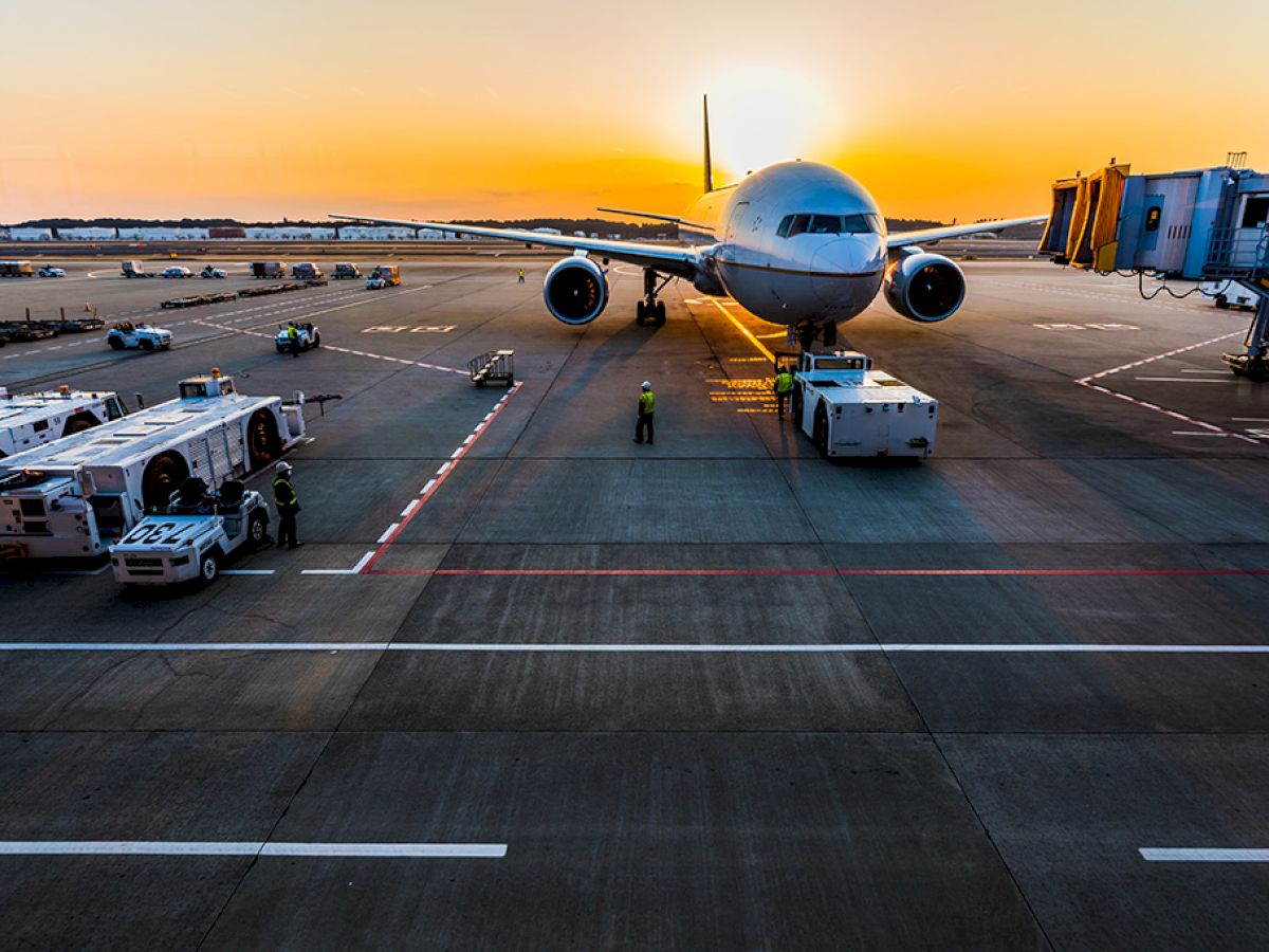 An airplane is parked at the gate of an airport at sunrise, with ground crew and vehicles around it, and the jet bridge connected.