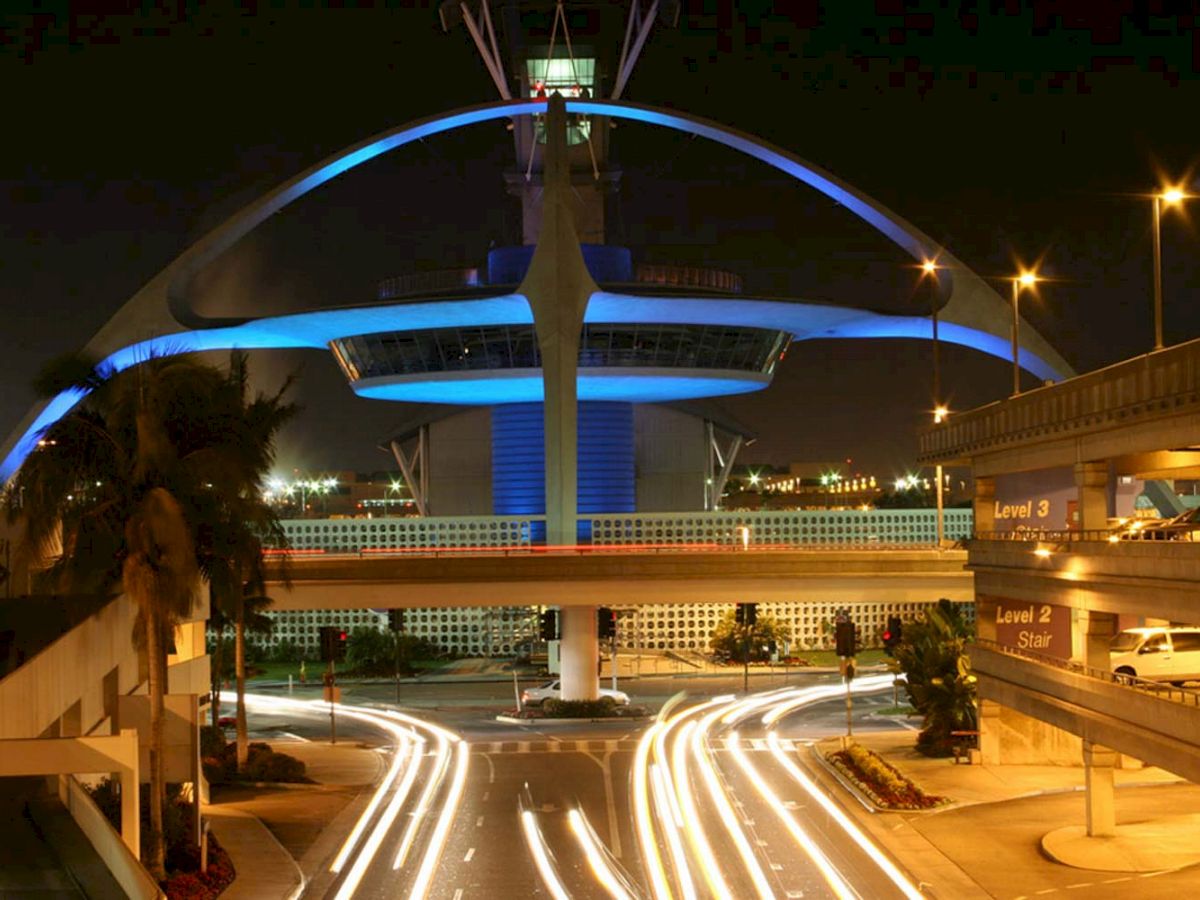 This image shows the iconic Theme Building at Los Angeles International Airport (LAX) at night, illuminated in blue light with light trails from cars.