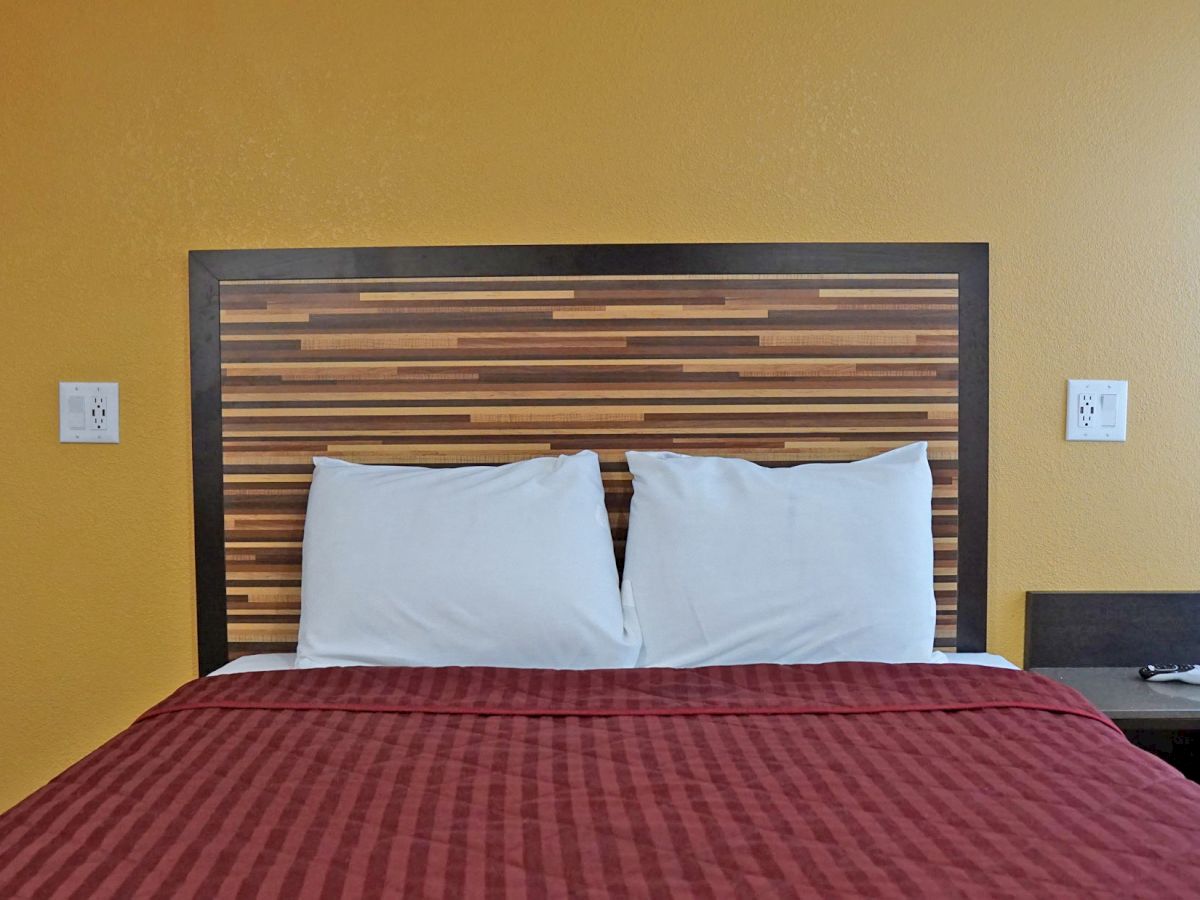 A neatly made bed with two white pillows, a red blanket, a wooden headboard, and a nightstand against a yellow wall, featuring two wall outlets.