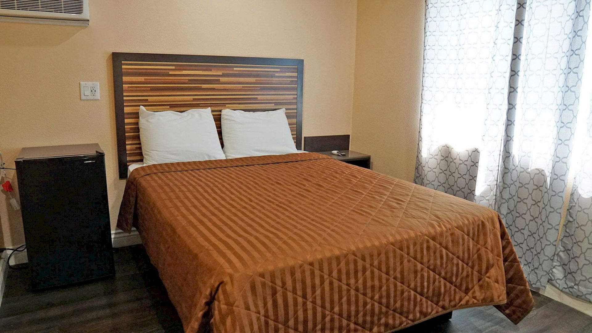 The image shows a neatly made bed with two pillows and a brown quilt in a room with beige walls, a nightstand, and a mini-fridge next to the bed.