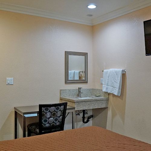 The image shows a small, beige hotel room with a bed, a wall-mounted TV, a desk and chair, a sink with a mirror, and a towel on a rack.