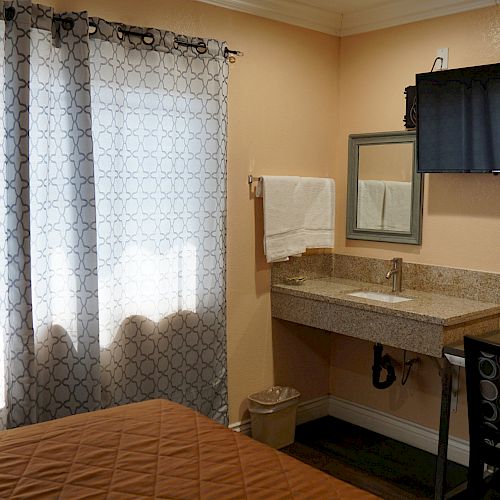 The image shows a small hotel room with a bed, window with curtains, wall-mounted TV, small desk, mirror, towel rack, and a countertop sink.