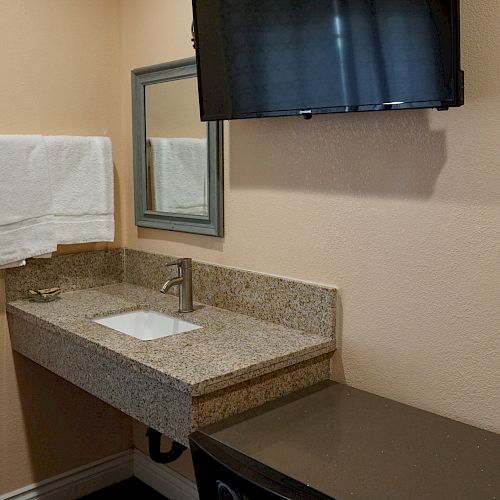 A bathroom with a granite countertop sink, a mounted towel, a wall mirror, and a TV above a black cabinet.