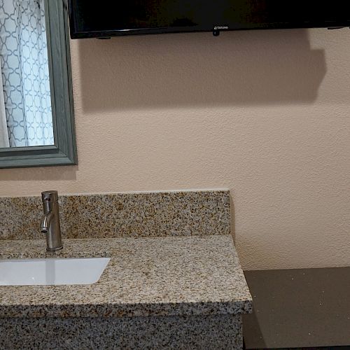 The image shows a small bathroom counter with a sink, a mirror above it, a wall-mounted television, and a towel hanging beside the mirror.