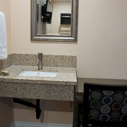 The image shows a small bathroom or vanity area with a sink, mirror, towel rack, and a desk with a chair beside it.