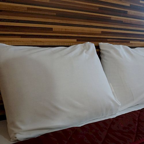 The image shows a neatly made bed with two white pillows and a maroon quilted bedspread against a wooden headboard.