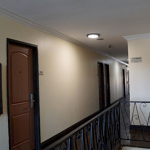 The image shows a hotel or apartment hallway with beige walls, numbered brown doors, a staircase railing, and a ceiling light fixture.