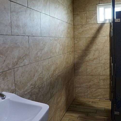 A clean, tiled restroom with beige walls, a white sink, and a stall door. A small window allows natural light to enter the room.