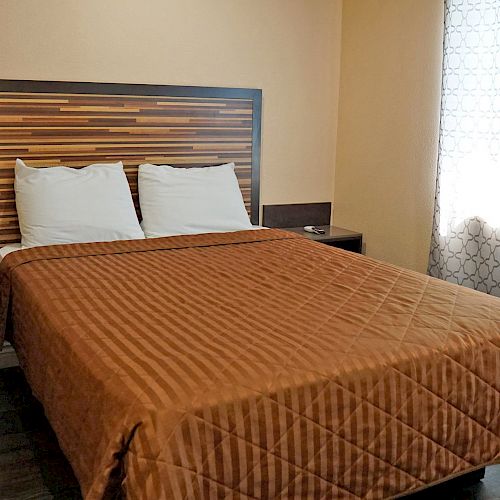 A neatly made bed with a brown quilt, two pillows, a mini-fridge, bedside table, decorative headboard, and window with patterned curtains.