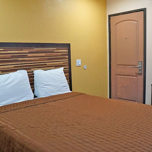 A small bedroom with a double bed, two white pillows, a wooden headboard, a door, and a chair at a desk, all under warm yellow lighting.