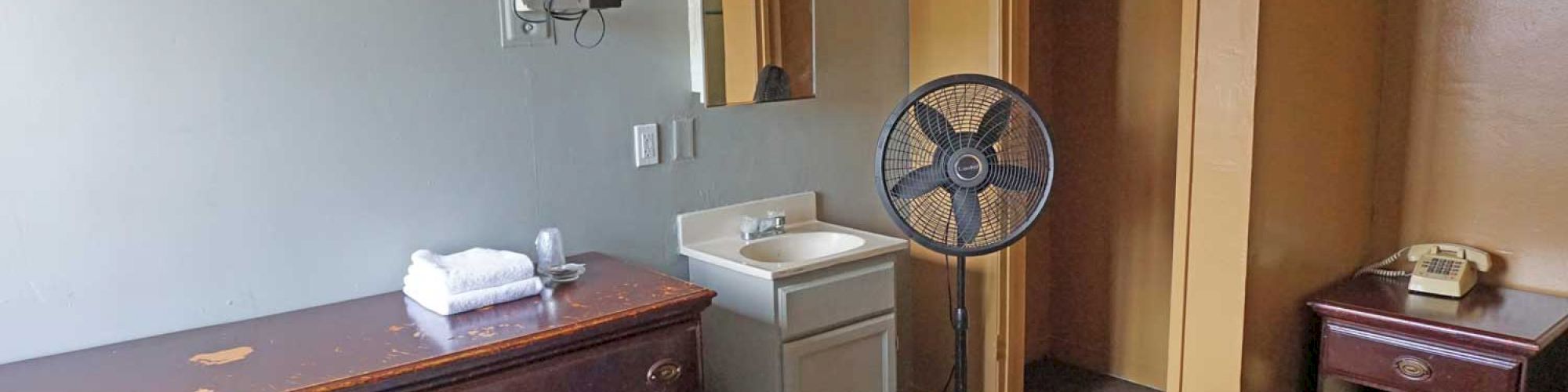 The image shows a basic room with a bed, dresser, wall-mounted TV, fan, sink, and an open closet. The bed has a multicolored quilted cover.