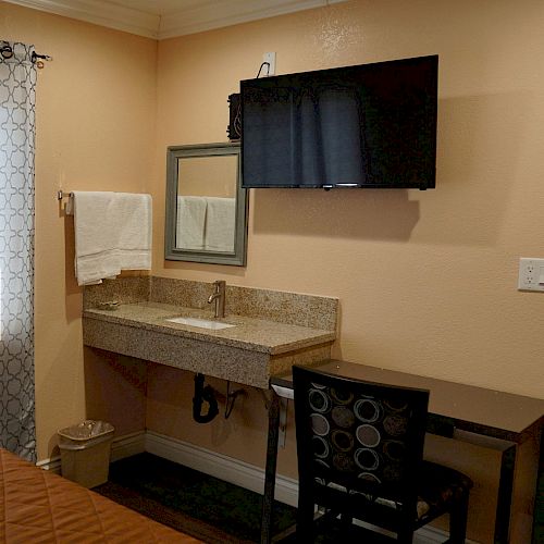 The image shows a hotel room with a bed, curtains, wall-mounted TV, a sink area with a mirror, a towel, a trash bin, and a desk with a chair.