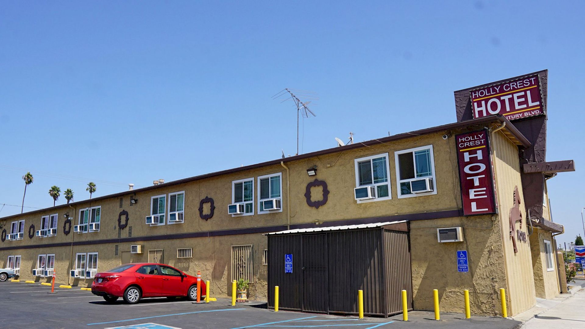 An image of a two-story hotel named "Holly Crest Hotel," with a red car parked in front and a clear blue sky in the background.