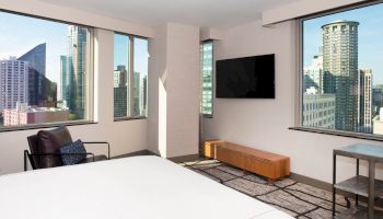 A modern hotel room with large windows showcasing an urban skyline, a bed, a TV, a chair, and a small table and chair.