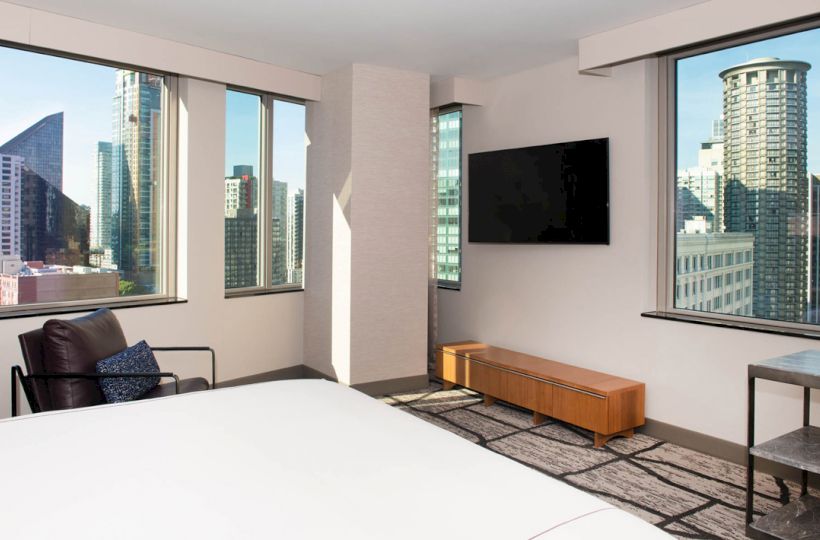 A modern hotel room with large windows showcasing an urban skyline, a bed, a TV, a chair, and a small table and chair.
