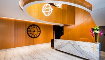 The image shows a modern interior with a marble reception desk, a large wall clock, wooden accents, and decorative lighting fixtures.
