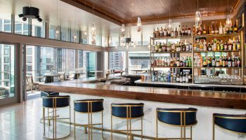 The image shows a modern bar with a wooden countertop, stylish bar stools, and a fully stocked shelf of various bottles, set against large windows.