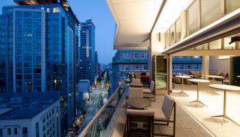 The image shows a modern balcony with tables and chairs, overlooking a city street lined with tall buildings at dusk.