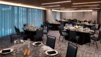 A banquet hall is set up with round tables covered with black tablecloths, glassware, plates, napkins, and floral centerpieces.