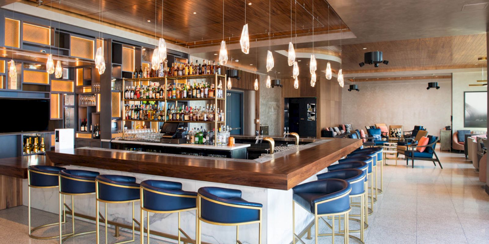 A modern bar with a wooden counter, blue bar stools, and hanging lights. Shelves filled with bottles are behind the counter.