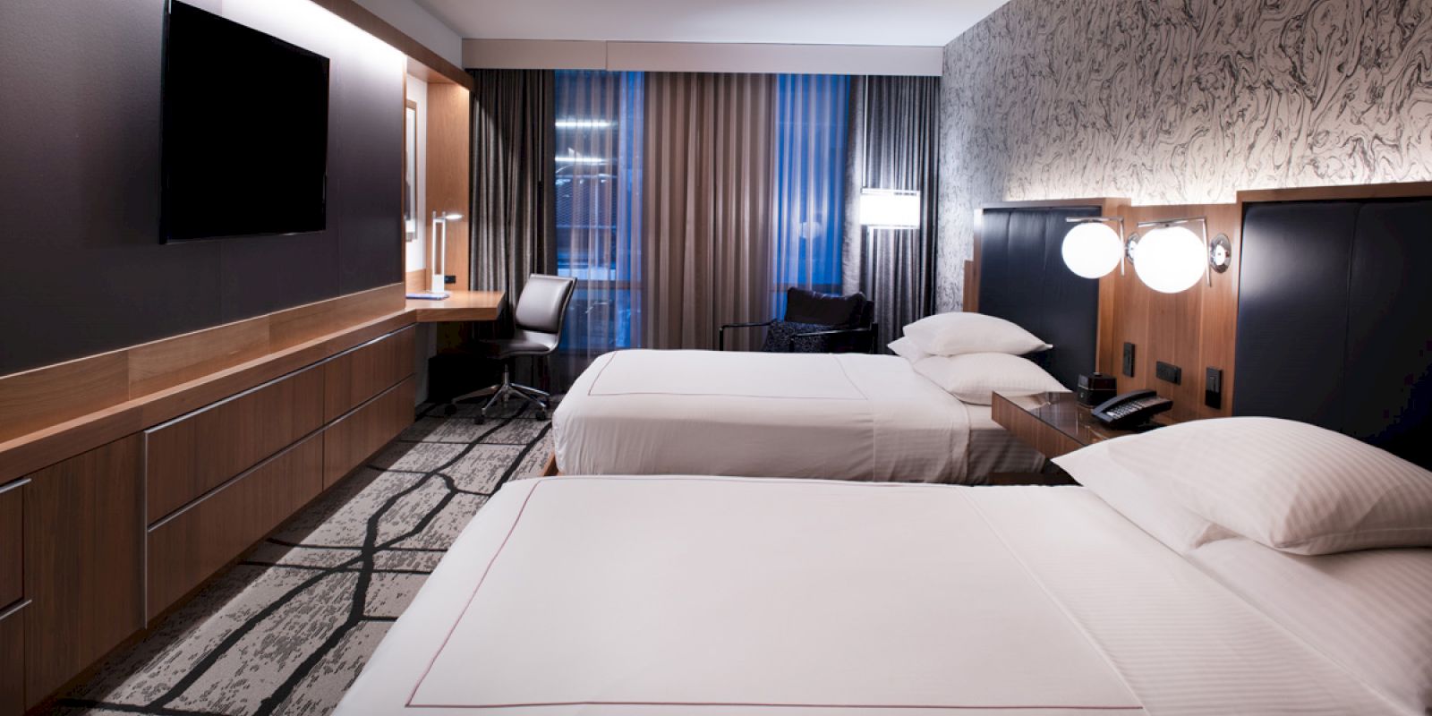 A modern hotel room with two neatly made twin beds, a desk with a chair, wall-mounted TV, and large windows with curtains, featuring stylish decor.