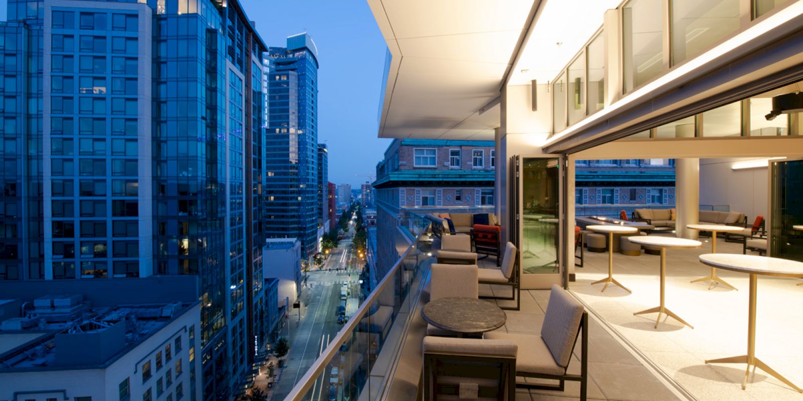 The image shows a modern urban balcony at dusk overlooking a cityscape with tall buildings and a busy street below, featuring outdoor seating areas.