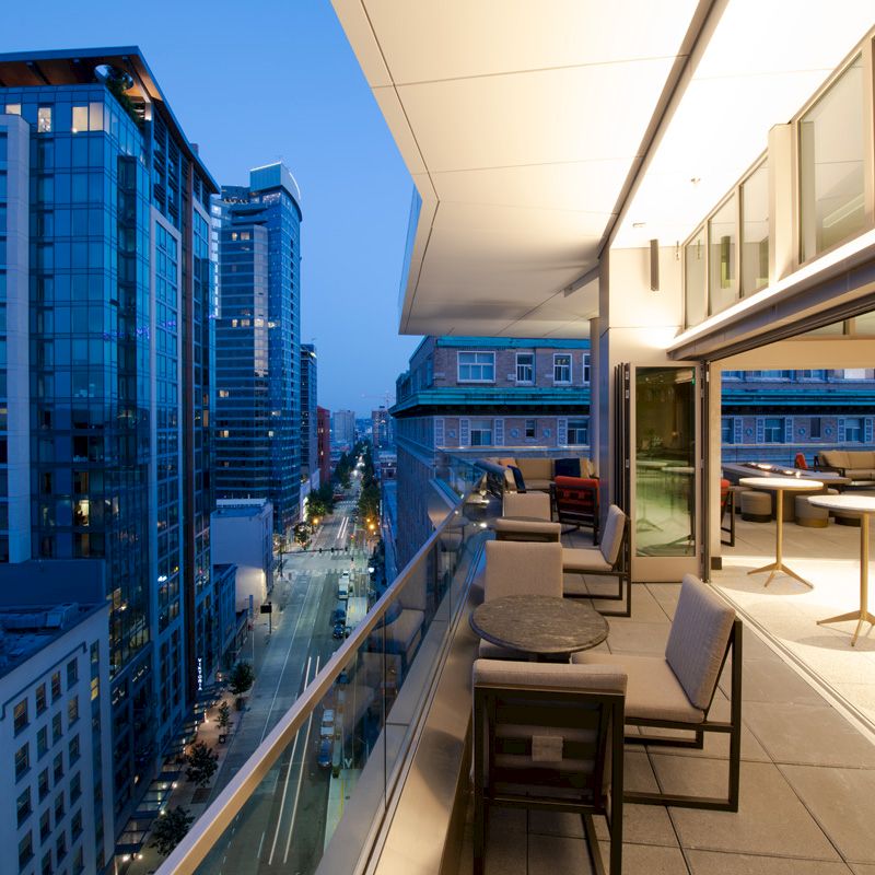A modern balcony with tables and chairs overlooks a city street lined with illuminated skyscrapers and buildings at dusk.