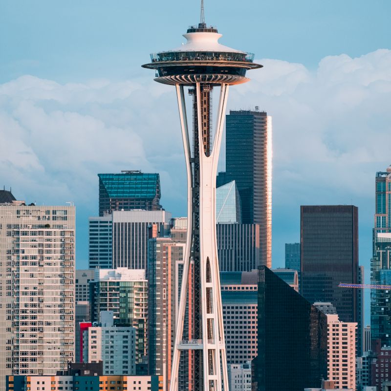 The image shows the Space Needle in Seattle, with a backdrop of various high-rise buildings and a partially cloudy sky in the background.