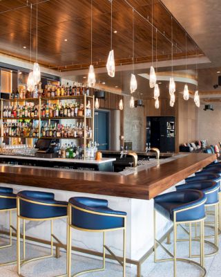 A modern bar with blue bar stools, a wooden countertop, hanging pendant lights, and a fully stocked liquor shelf in a stylish and spacious lounge.