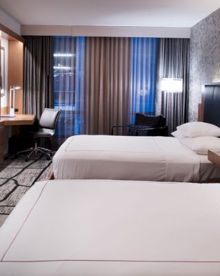 A modern hotel room with two double beds, a wall-mounted TV, a desk with a chair, and contemporary decor featuring patterned wallpaper and carpeting.