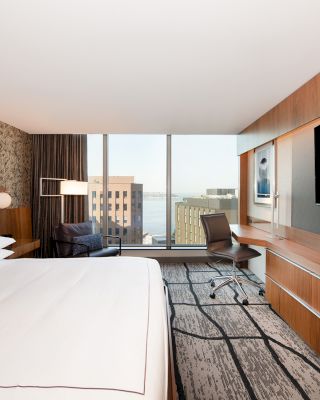 A modern hotel room with a large bed, wall-mounted TV, desk, and a view of city buildings through floor-to-ceiling windows ends the sentence.