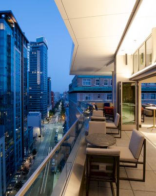 A modern balcony overlooks a city street lined with tall buildings. There are tables and chairs on the balcony for seating.