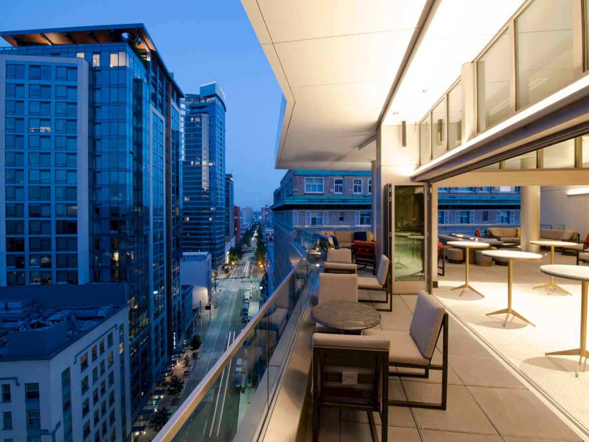 A modern cityscape view from a balcony at dusk, featuring outdoor seating and tables, with tall buildings and a street seen below.