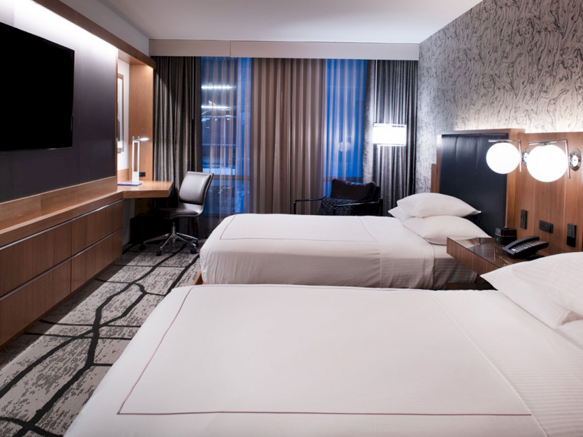 A modern hotel room with two neatly made beds, a wall-mounted TV, a work desk, and a large window with curtains and soft lighting ends the sentence.