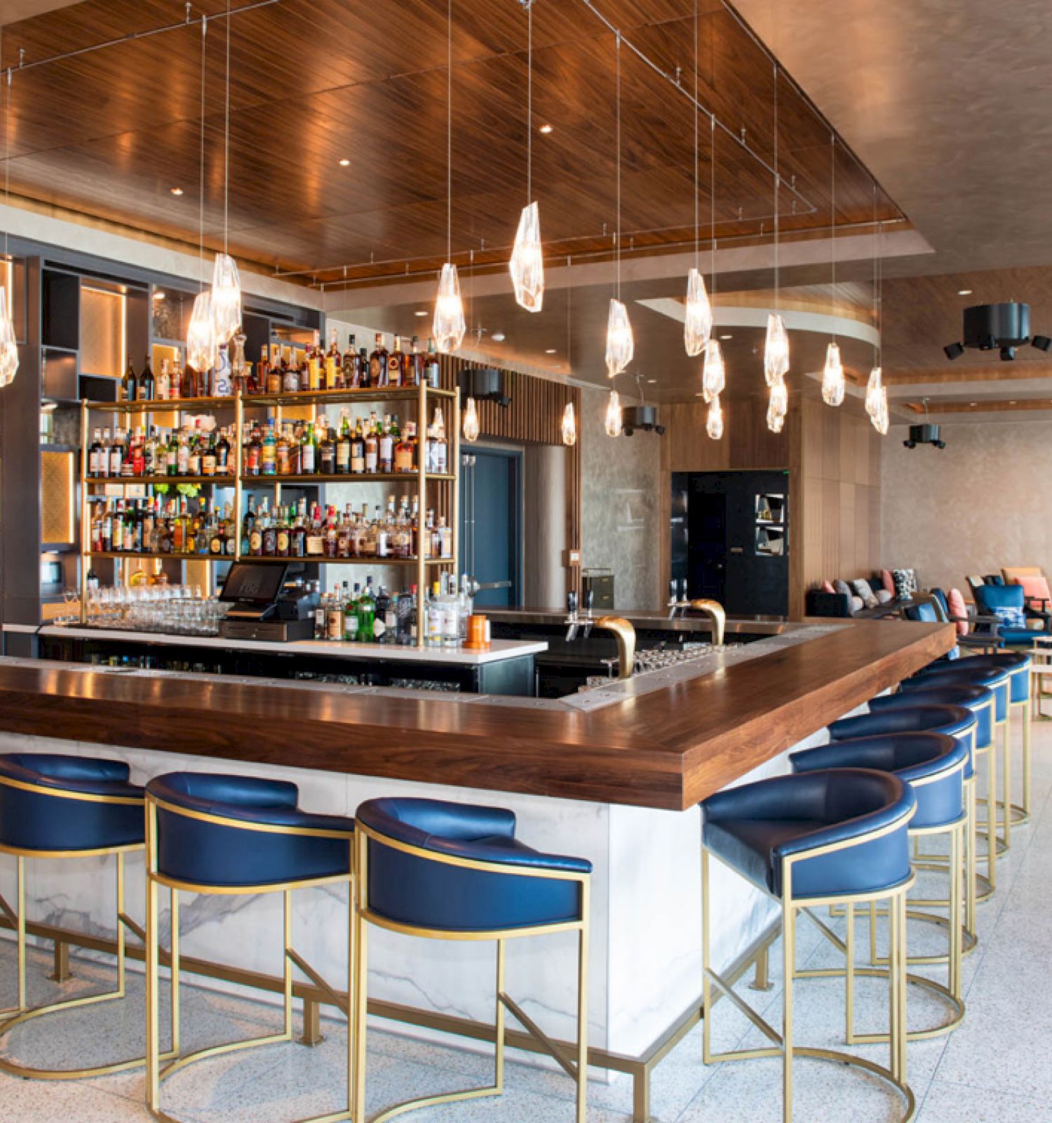 A modern bar with a well-stocked shelf, stylish lighting fixtures, blue cushioned bar stools, and a comfortable seating area in the background.