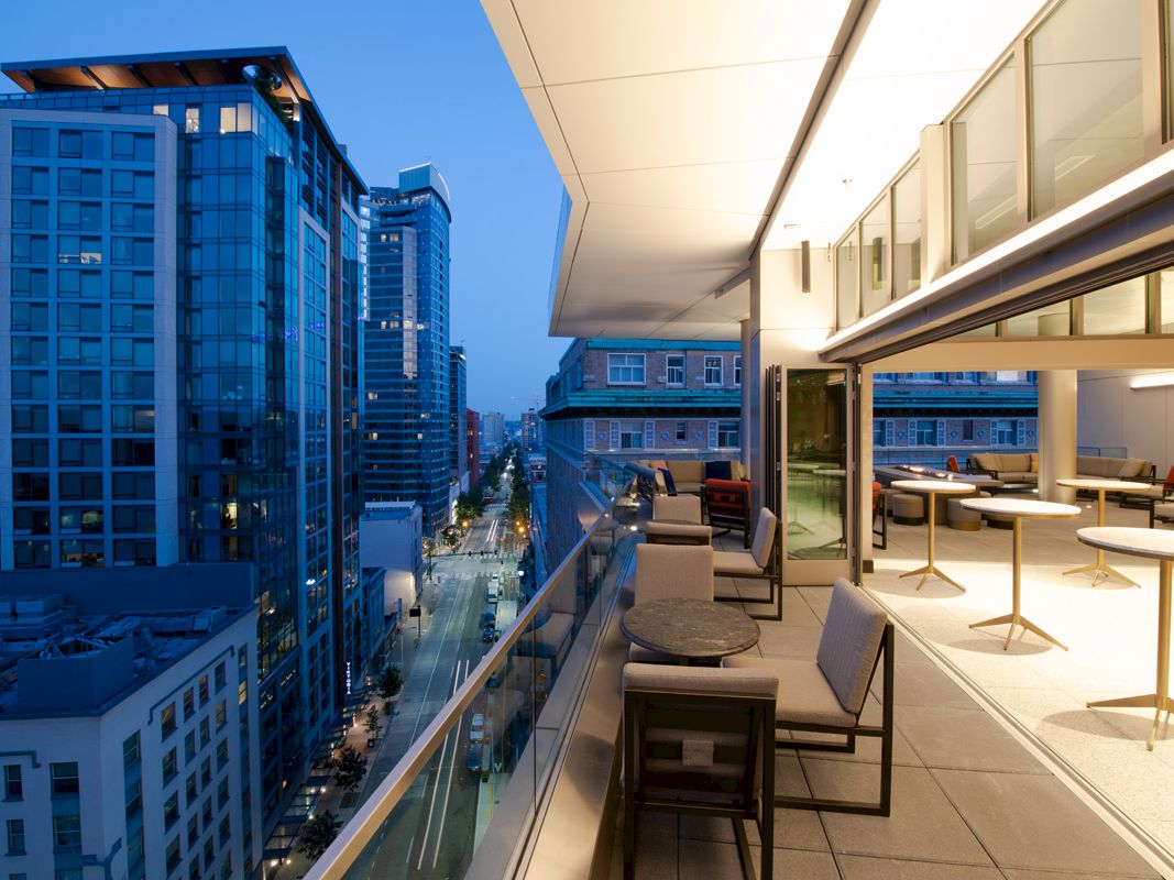 A modern outdoor terrace with seating overlooking a city street lined with tall buildings during the evening.