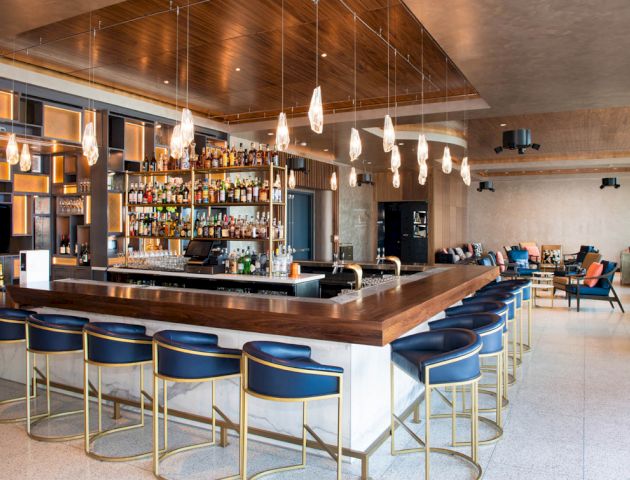 A modern bar with sleek blue and gold bar stools, a well-stocked shelf, overhead lighting, and a seating area with chairs and tables in the background.