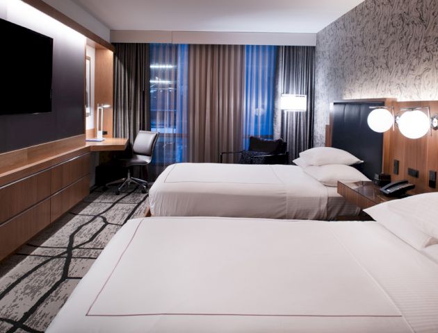 The image shows a modern hotel room with two neatly made beds, a flat-screen TV, a desk, a chair, and stylish lighting.