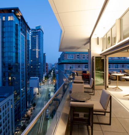 A modern cityscape at dusk shows tall buildings and a balcony with chairs and tables, overlooking a busy street below.