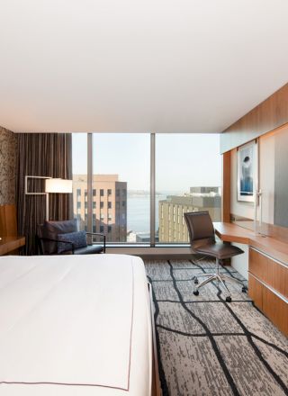 A modern hotel room with a large bed, flat-screen TV, desk, chair, and a window offering a cityscape view.