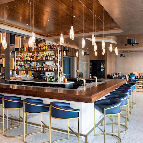 The image displays a modern bar with blue stools, a well-stocked bar area, hanging pendant lights, and a seating area with orange and blue chairs.