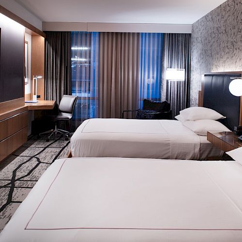 A modern hotel room with two neatly made beds, a desk and chair by the window, a wall-mounted TV, and minimalistic decor.