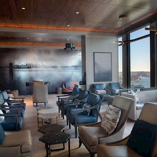 A modern lounge with comfortable seating, large windows displaying a waterfront view, and a decorative wall mural of a skyline.