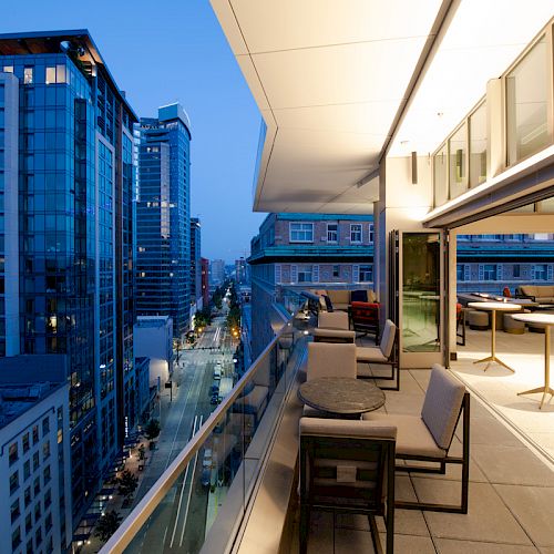 A modern balcony with tables and chairs overlooks a city street lined with tall buildings at dusk, showcasing a sophisticated urban setting.