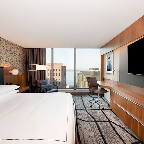 Modern hotel room with large bed, wall-mounted TV, desk, chair, and a view of city buildings through floor-to-ceiling windows.