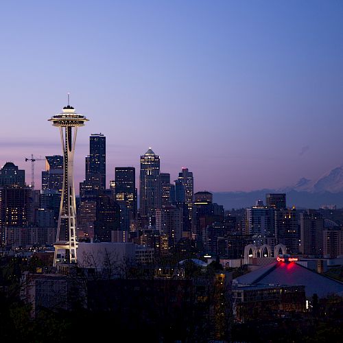 This image shows the Seattle skyline at dusk, featuring the Space Needle prominently and Mount Rainier in the background, against a purple-blue sky.