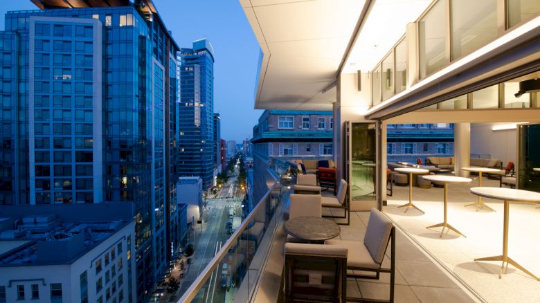 A modern outdoor balcony with tables and chairs overlooking a city street lined with high-rise buildings at dusk, creating a vibrant urban scene.