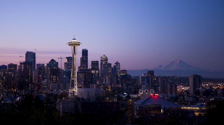 The image depicts the Seattle skyline at dusk, featuring the Space Needle prominently, with Mount Rainier visible in the background.