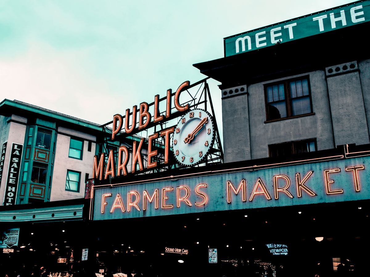 This image shows the iconic neon signs of a Public Market and a Farmers Market, featuring a clock. The signs are mounted on buildings in an urban setting.