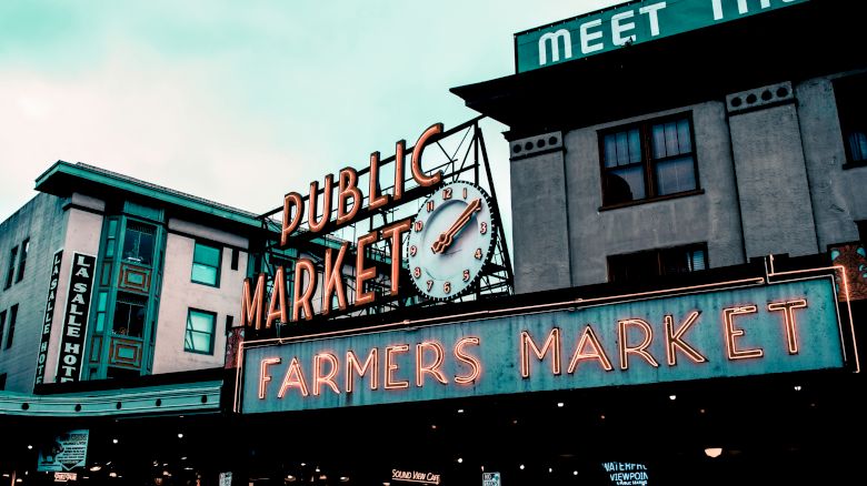 This image shows the iconic neon signs of a Public Market and a Farmers Market, featuring a clock. The signs are mounted on buildings in an urban setting.